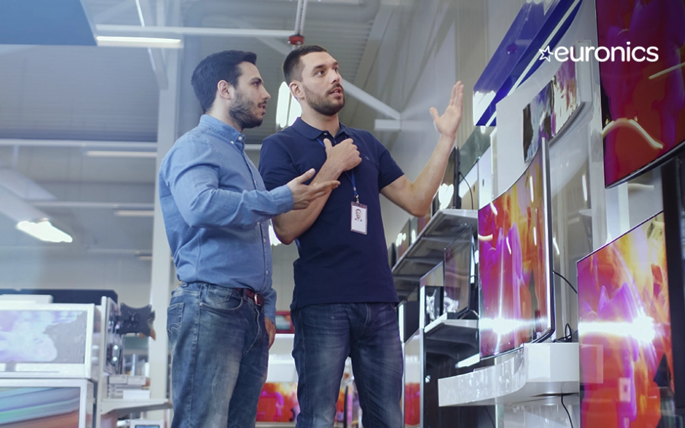 zwei Männer unterhalten sich und schauen auf Fernseher im Technikladen. In der rechten oberen Ecke das Logo des NETFORMIC Kunden Euronics
