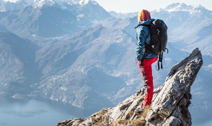 Eine Person in Wanderkleidung und mit Rucksack steht auf der Klippe und schaut in ferne auf die Berge.