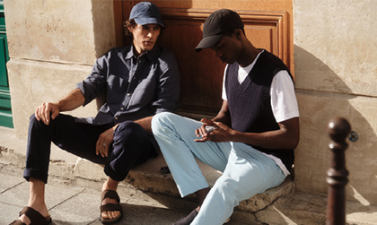 Zwei Männer in Modekleidung unterhalten sich sitzend draußen vor einer Haustür. 