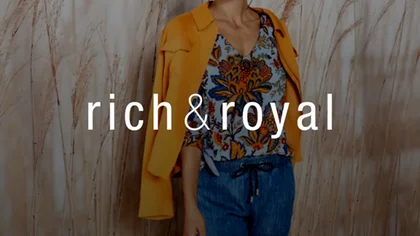 Frau gekleidet in Jeans, gemusterten Bluse, Jacke in orange steht vor einer Wand mit Fototapete mit Schilf. Darüber das Logo des NETFORMIC Kunden Rich & Royal