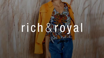 Frau gekleidet in Jeans, gemusterten Bluse, Jacke in orange steht vor einer Wand mit Fototapete mit Schilf. Darüber das Logo des NETFORMIC Kunden Rich & Royal