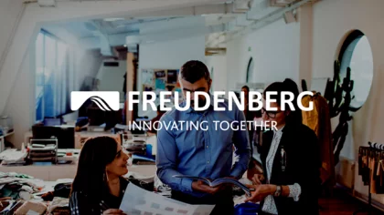 NETFORMIC Kundencase Freudenberg Menschen mit Magazinen und Unterlagen unterhalten sich im Büro. Darüber das Logo des NETFORMIC Kunden Freudenberg