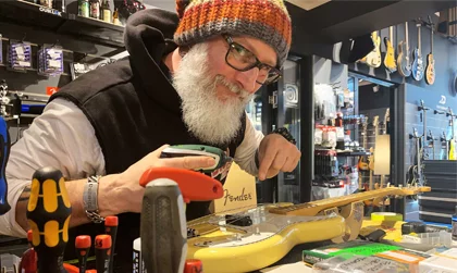 Session Kundencase Ein weißbärtiger Mann mit einer farbigen Mütze und einer Brille steht an der Theke in einem Musikinstrumenteladen, schaut in die Kamera und zeigt auf eine Gelbe Gitarre vor ihm.